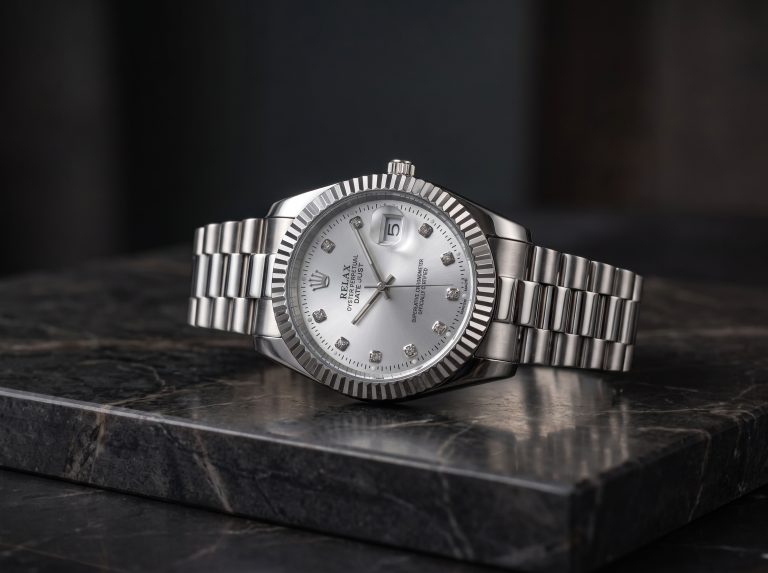 Silver luxury men's wristwatch with a silver dial, diamond-style hour markers, fluted bezel, date display, and stainless steel bracelet on a black marble surface.